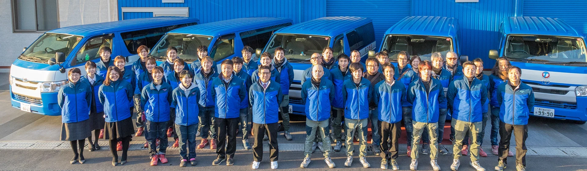 青い社屋と車両の前に並ぶ株式会社つきののスタッフ